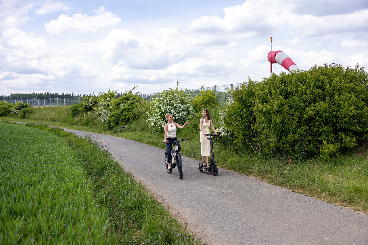 Das Bild zeigt 2 Damen welche eine mit einem E-Roller und die andere mit einem E-Bike am Flughafen Hof-Plauen fahren